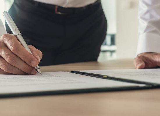 Businessman signing contract, document or report in a binder while standing at his office desk.