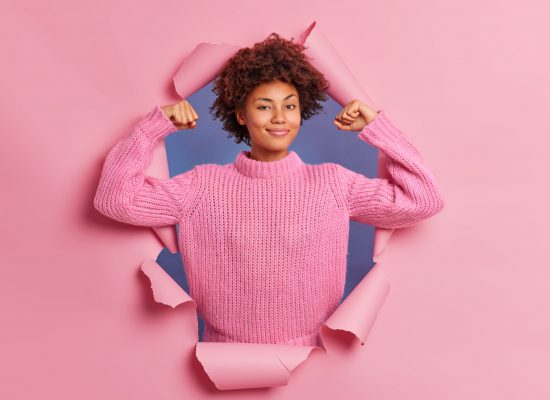 Self assured beautiful young Afro American woman raises arms shows biceps being strong and powerful proud of her own achievements wears casual knitted sweater breaks through paper background